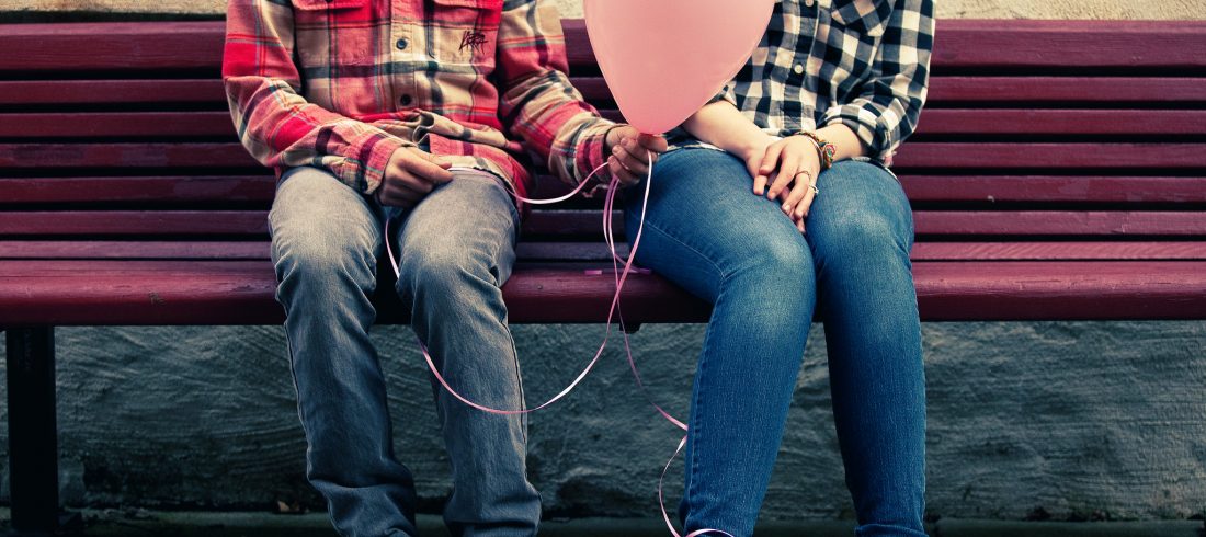 Two people sitting on a bench, each with shy body language, one offering to give the other a balloon