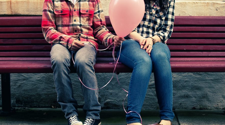Two people sitting on a bench, each with shy body language, one offering to give the other a balloon