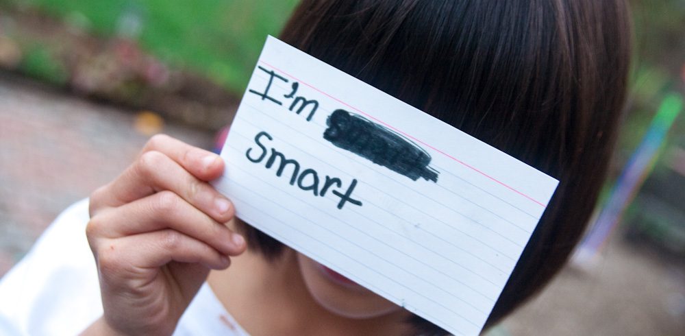 a child holds a notecard in front of her face; the notecard reads “I’m smart” in black marker. “Smart” was written once, scratched out, written again.