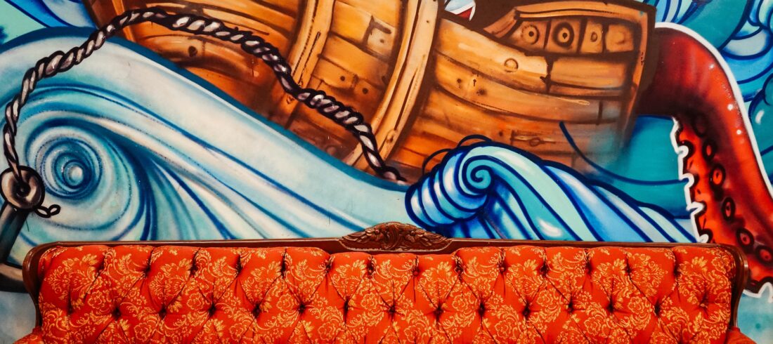On the wall, a mural depicts a boat tossed by waves, struggling to lay anchor. In the front, a bold red couch rests comfortably, welcoming contemplation.
