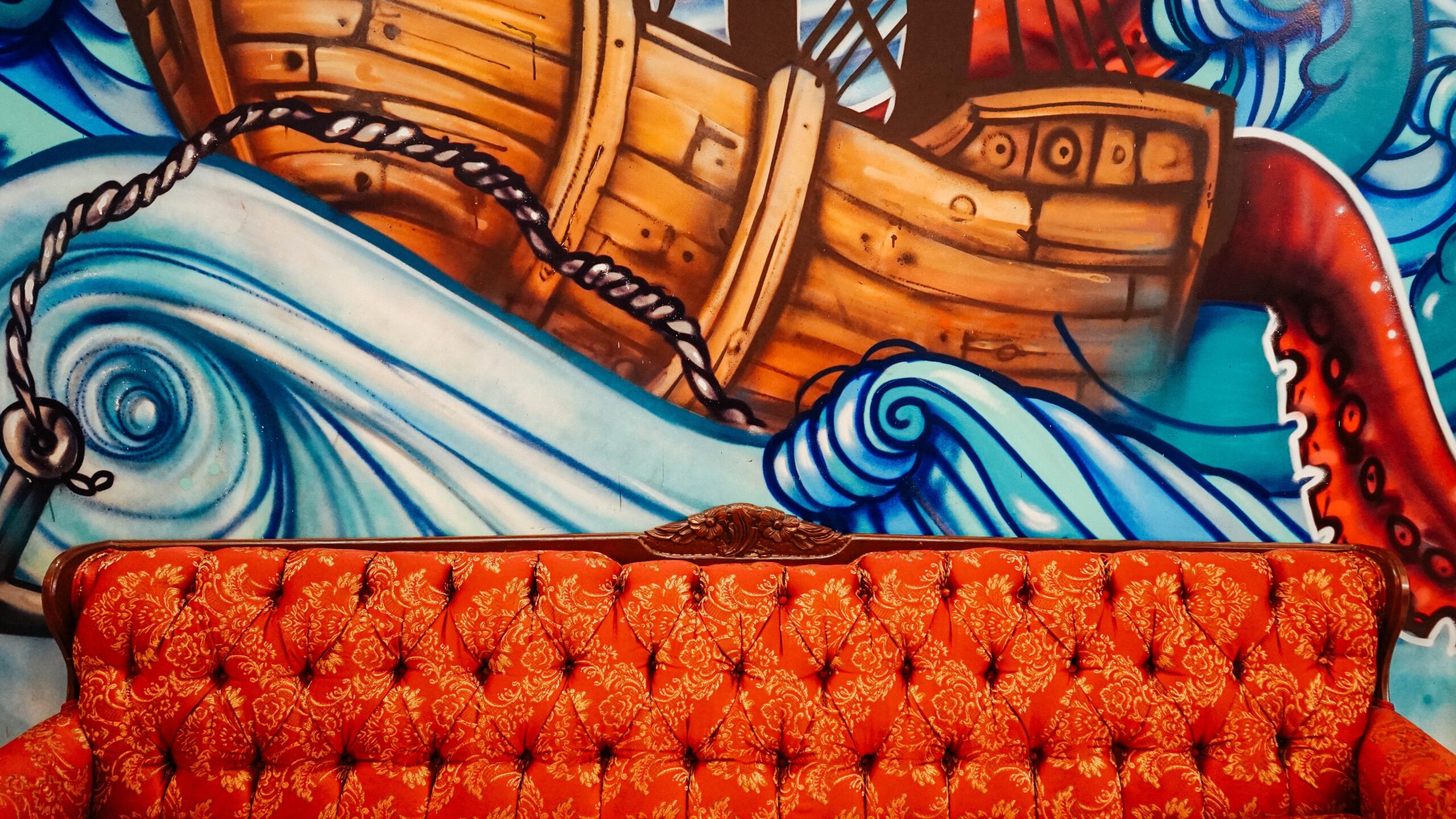 On the wall, a mural depicts a boat tossed by waves, struggling to lay anchor. In the front, a bold red couch rests comfortably, welcoming contemplation.