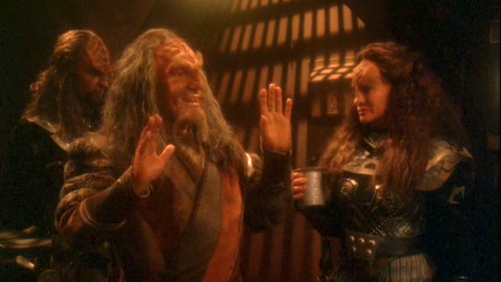 Still taken from Star Trek episode, showing three Klingons in a dimly lit room. Character in foreground is smiling, accepting but downplaying acclaim from others off-screen.