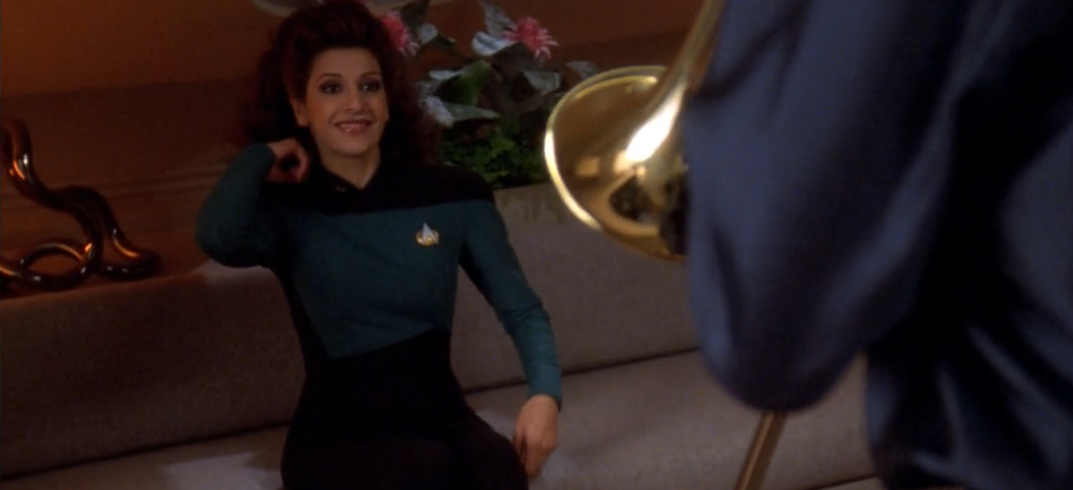 dark-haired woman in uniform smiles at the man (off-camera) playing trombone nearby