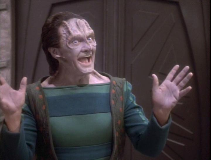 A wide-grinned alien in shirt and vest raises jazz hands to get you excited; he's probably trying to sell you something. Wait, it's Garak. He's definitely trying to sell you something.