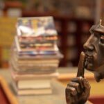A small statue of a shushing finger in front of a face rests on a shelf. In the background, stacks of books. What information do we lose by removing sound from our texts?